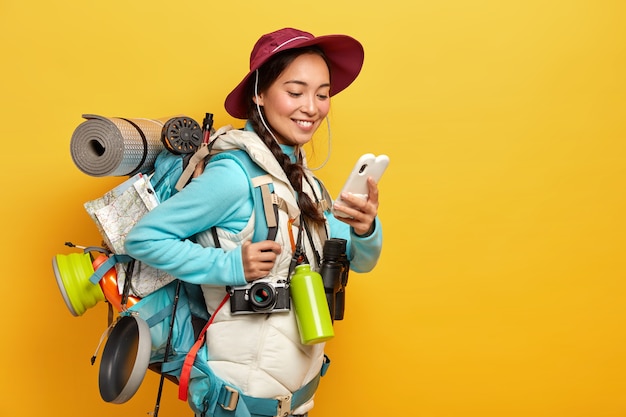 Positieve mooie aziatische toerist gebruikt moderne gadget om te navigeren, gekleed in hoed en trui met jas, verrekijker, retro camera, karemat gebruikt tijdens reis staat tegen gele muur