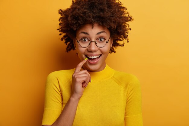 Positieve nieuwsgierige vrouw luistert naar iets heel interessants, houdt de vinger op de lippen, heeft een directe blik, praat graag over aangenaam nieuws, draagt een bril, heeft ogen vol interesse en geluk