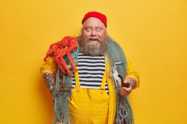 Positieve onder de indruk bebaarde matroos in gestreept vest vormt met rode octopus op schouder, rookpijp houdt, draagt visnet