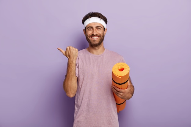 Positieve ongeschoren man draagt witte hoofdband en paars t-shirt, houdt opgerolde mat vast, wijst opzij