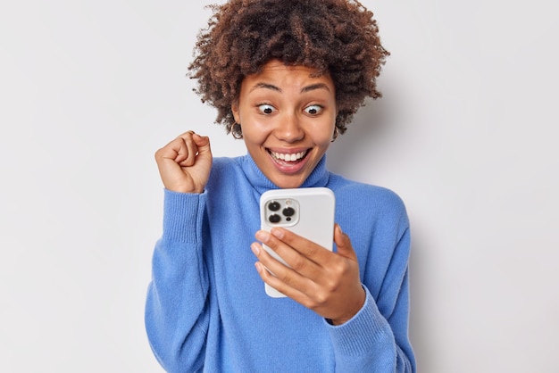 Positieve optimistische jonge vrouw balt vuist kijkt met opwinding naar smartphonescherm reageert op geweldig nieuws draagt blauwe coltrui geïsoleerd op witte achtergrond ontdekt dat ze de loterij heeft gewonnen