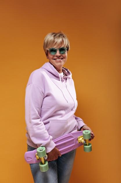 Positieve oude dame met blond haar en trendy zonnebril in roze modieus sweatshirt en coole spijkerbroek glimlachend en skateboard te houden.