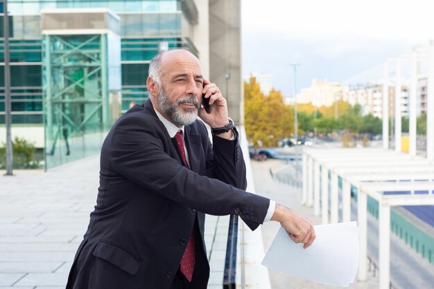 Positieve peinzende grijze haired zakenman die op cellphone spreekt