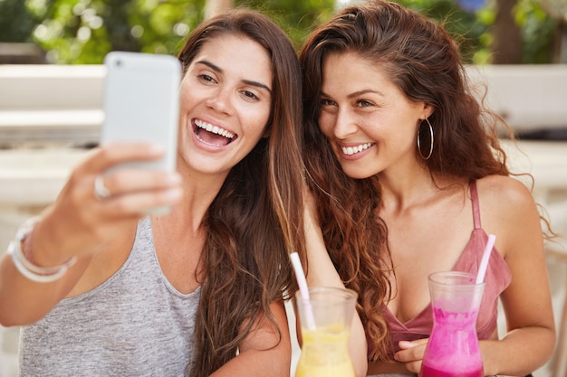 Positieve, prachtige vrouwen die in een goed humeur zijn, genieten van recreatietijd op een gezellige terrasje, poseren voor selfie, drinken een verse smoothie, hebben samen plezier.