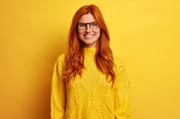 Positieve roodharige Europese vrouw met een blij gezicht glimlacht aangenaam voelt zich gelukkig na een succesvolle dag draagt een warme gele trui.