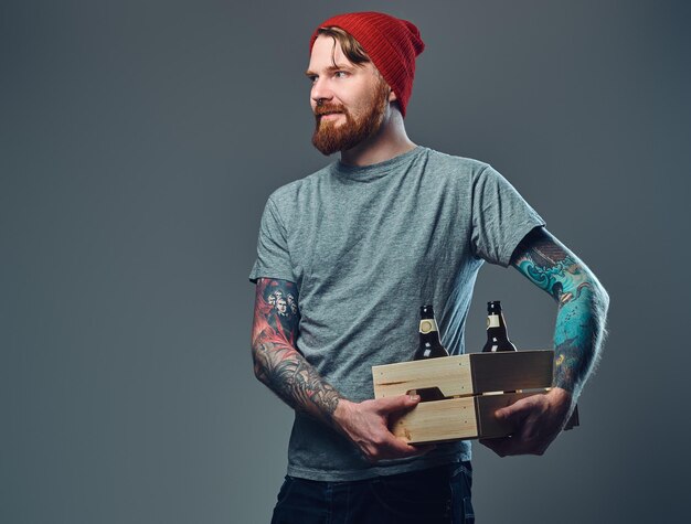 Gratis foto positieve roodharige getatoeëerde, bebaarde man houdt een houten kist met bierflesjes over grijze achtergrond.