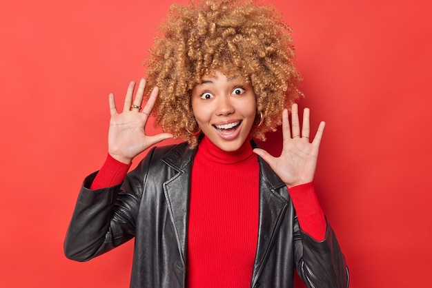 Positieve speelse vrouw met krullend haar houdt handpalmen omhoog probeert je te vermaken glimlacht breed heeft plezier draagt casual poloneck en leren jas geïsoleerd over levendige rode achtergrond geluk concept