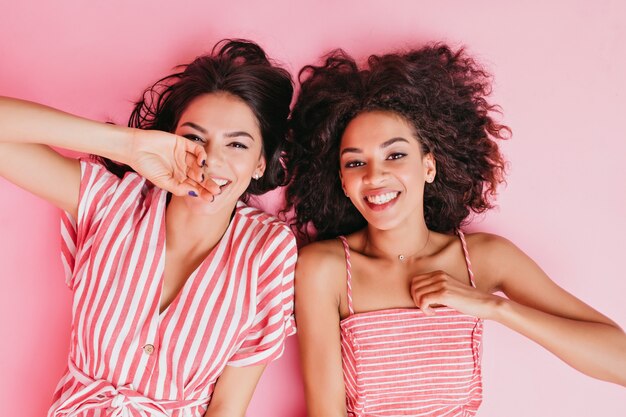 Positieve vriendinnen liggen te genieten van rust in de kamer van het roze meisje. Portret van gebruinde brunettes in gestreepte overgooiers.