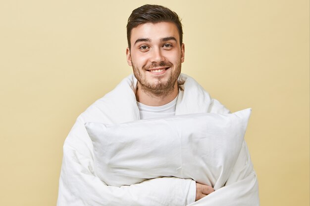 Positieve vrolijke jongeman met schattige glimlach en ongeschoren gezicht staande op een blinde muur, gewikkeld in een witte deken, zich dolgelukkig voelen, herstellende van de kou, kussen vasthoudend, in bed gaan slapen