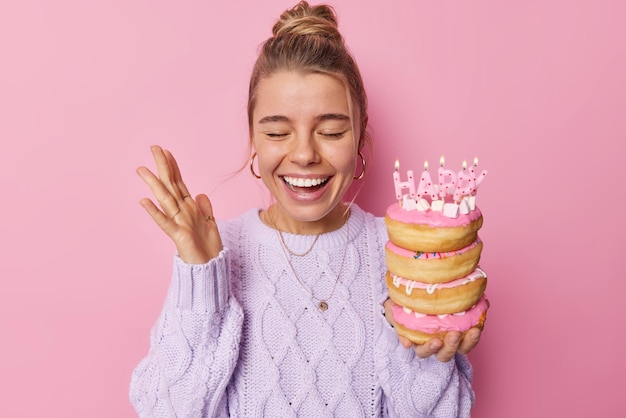 Positieve vrouw geniet van verjaardagsviering houdt palm omhoog gegiecheld houdt gelukkig stapel heerlijke donuts met kaarsen draagt casual trui geïsoleerd over roze achtergrond. feestelijke gelegenheid