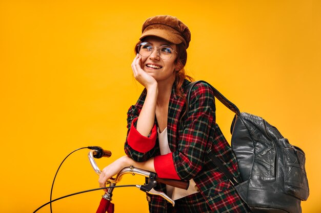 Positieve vrouw in fluwelen pet en bril poseert dromerig met fiets with