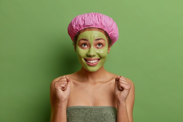 Positieve vrouw kijkt vrolijk boven gebalde vuisten wacht op iets speciaals draagt badmuts en handdoek rond lichaam past voedend avocado-masker toe op gezicht geïsoleerd op groene muur