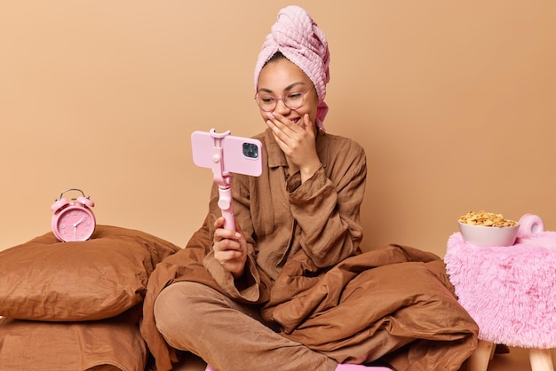 Positieve vrouw lacht vrolijk houdt hand op mond poses voor smartphone neemt video op of maakt online oproep draagt pyjama en handdoek gewikkeld op hoofd poses op comfortabel bed Goedemorgen