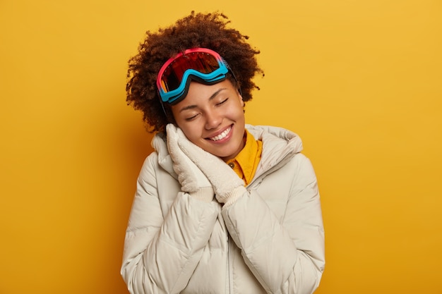 Positieve vrouw met Afro-kapsel leunt tegen geperste handpalmen, heeft een dromerige gezichtsuitdrukking, draagt een witte jas, handschoenen en een snowboardmasker, geniet van winteravontuur, geïsoleerd op een gele muur.