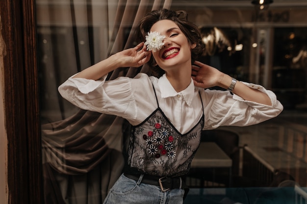 Positieve vrouw met kort haar in wit en zwart shirt met lange mouwen glimlachend oprecht in café. vrolijke dame die in spijkerbroek bloem binnen houdt.