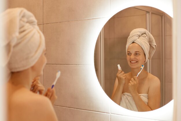 Positieve vrouw met witte handdoek op haar hoofd met tandpasta en tandenborstel in handen, kijkend naar haar reflectie in de spiegel met een gelukkige gezichtsuitdrukking.