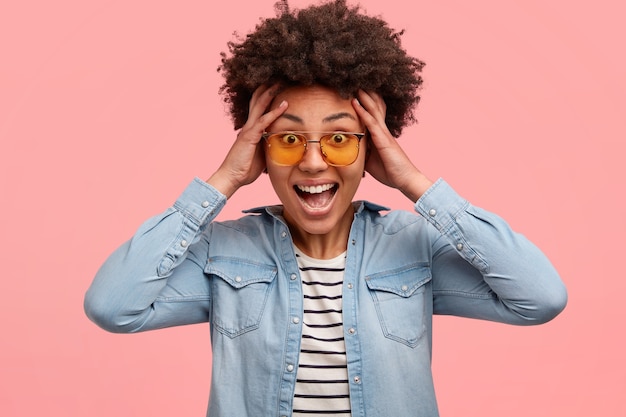 Positieve vrouw van gemengd ras houdt de handen op het hoofd, heeft een afro-kapsel, ontvangt een ongelooflijke verrassing