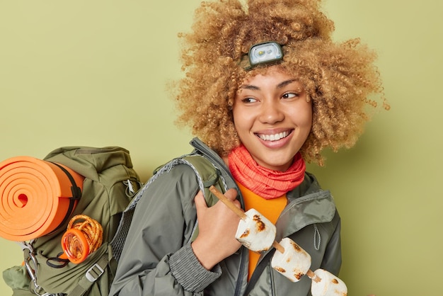 Positieve vrouwelijke camper heeft picknick houdt geroosterde marshmallow bereidt zich voor op barbecue draagt headmalp en jas draagt zware rugzak geïsoleerd over donkergroene achtergrond Toerisme en camping concept