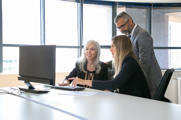 Positieve zakencollega's kijken naar presentatie op pc-monitor, project bespreken, zitten op de werkplek en wijzen op display. Zakelijke communicatie concept