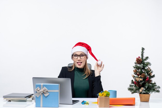 Positieve zakenvrouw spelen met haar kerstman hoed zittend aan een tafel met een kerstboom en een cadeau erop en tong uitsteken op witte achtergrond