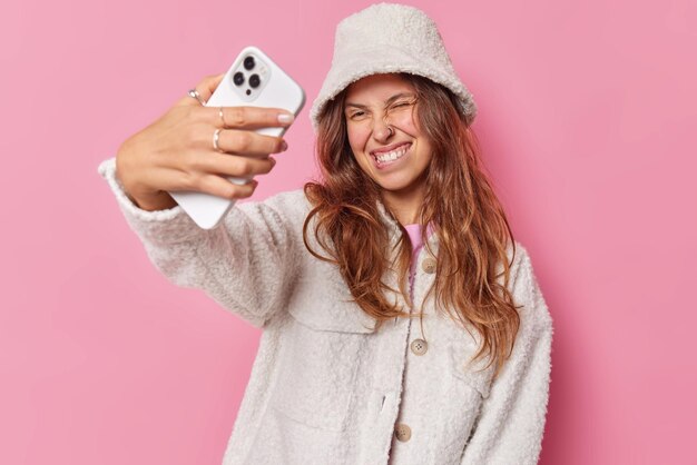 Positieve zorgeloze optimistische vrouw knipoogt oog bijt lippen en poseert op smartphone camera neemt selfie draagt modieuze witte jas bontmuts klikken om foto van zichzelf geïsoleerd over roze achtergrond te maken