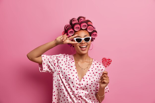 Positieve zorgeloze vrouw met donkere huid in trendy zonnebril maakt vredesgebaar over het oog, lacht vrolijk, heeft plezier, houdt lekkere lolly vast, draagt haarrollers voor het maken van perfecte krullen, nonchalant gekleed