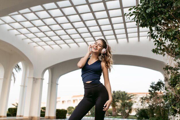 Positiviteit van vrolijke mooie vrouw in sportkleding uitdrukken door glimlachend op oost-architectuur te trainen. muziek luisteren via koptelefoons, geluk, sportieve levensstijl