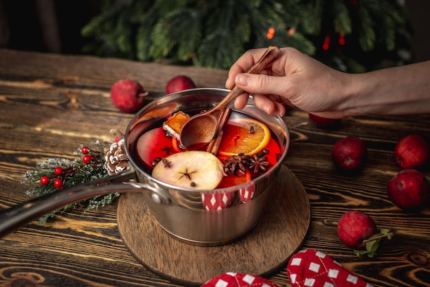 Pot met glühwein op een houten tafel met fruit en een kerstboom op de achtergrond. vrouw hand roert de drank met een lepel. concept van een gezellige feestelijke sfeer, nieuwjaarsstemming.