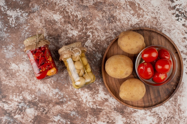 Potten van ingemaakte paprika's en champignons en plaat van gekookte aardappelen, ingelegde tomaten op marmeren oppervlak.