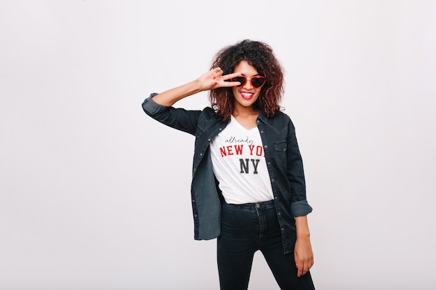 Prachtig Afrikaans vrouwelijk model in trendy t-shirt poseren met vredesteken en lachen. Verfijnd zwart meisje in rode zonnebril die voor de gek houdt