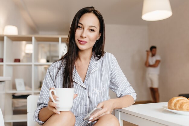 Prachtig brunette vrouwelijk model in blauw shirt poseren in keuken met kopje koffie