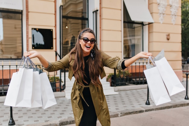 Gratis foto prachtig donkerharig vrouwelijk model met plezier op straat en zwaaiende winkeltassen