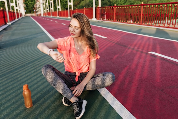 Prachtig Europees meisje in zwarte sneakers kijken naar haar polshorloge. Outdoor Portret van schattige jonge vrouw die zich voordeed op sintelbaan met SAP.