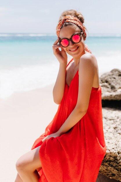 Prachtig gelooid meisje in zomerjurk poseren op het strand. Buiten schot van extatische lachende vrouw zittend op grote steen op het strand.