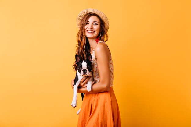 Prachtig gember meisje in zomer outfit met franse bulldog. Aantrekkelijke jonge dame in hoed lachen tijdens portretfotografie met puppy.