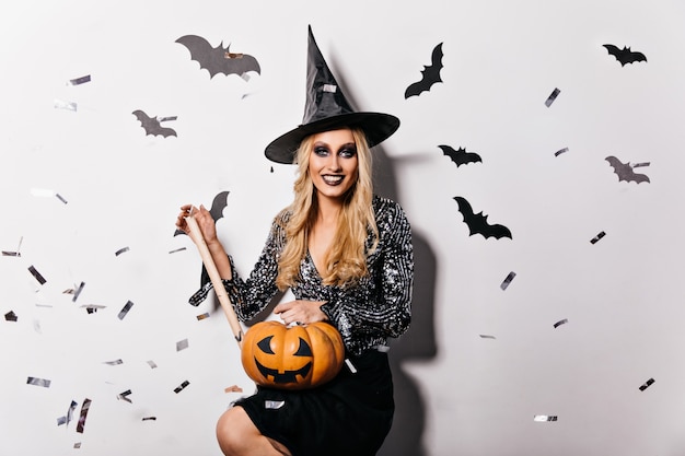 Prachtig meisje dat in fonkelingsblouse Halloween-pompoen houdt. Binnenfoto van glimlachende tevreden heks in zwarte hoed.