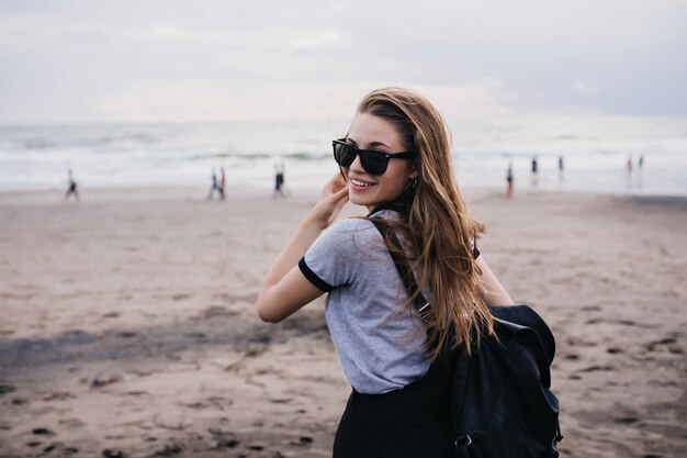 Prachtig meisje in zwarte zonnebril kijken over de schouder terwijl je aan zandstrand. Foto van mooi vrouwelijk model met blackpack poseren op aard.