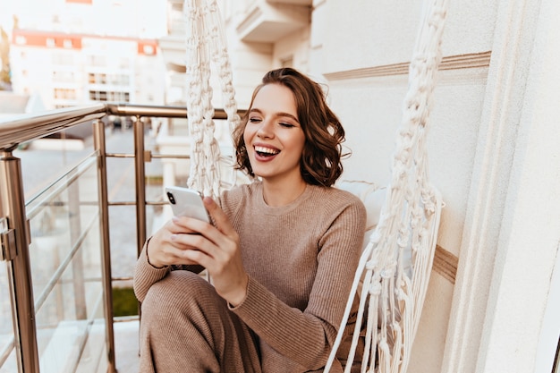 Prachtig meisje telefoonbericht lezen zittend op het terras. Foto van opgewonden schattige dame poseren met smartphone op balkon.