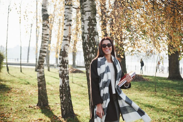 Prachtig model. De jonge het glimlachen brunette in zonnebril bevindt zich in het park dichtbij de bomen en houdt blocnote