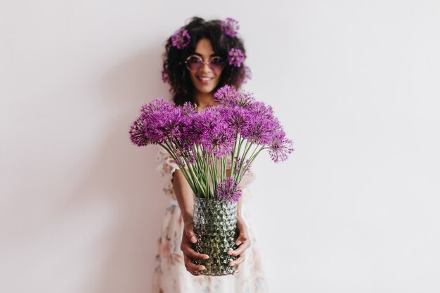 Prachtige Afrikaanse dame met vaas met paarse bloemen. Indoor portret van romantische zwarte jonge vrouw plezier thuis in weekend.