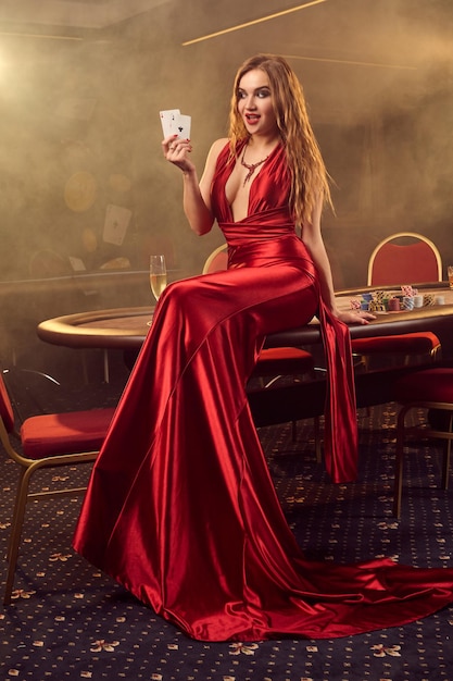 Prachtige blonde vrouw in een lange rode satijnen jurk, met twee azen in haar hand poseert zittend op een pokertafel in luxe casino. Passie, kaarten, chips, alcohol, winnen, gokken - het is een vrouwelijk vermaak