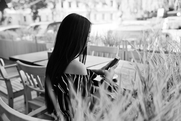 Prachtige brunette meisje zittend op de tafel in café met kopje koffie en het gebruik van telefoon