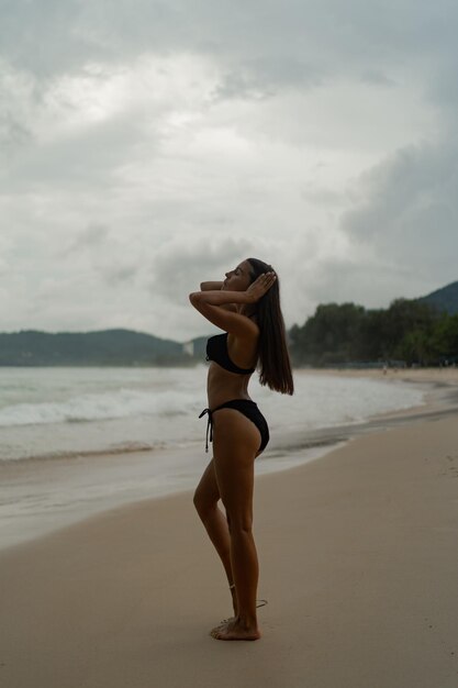 Prachtige brunette vrouw met perfect figuur poseren op tropisch strand, gekleed in stijlvolle zwarte badmode