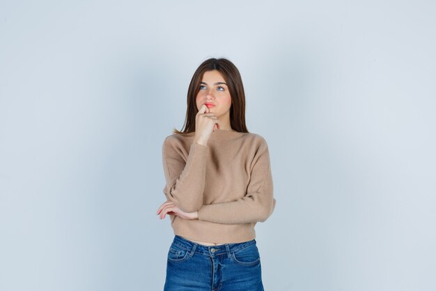 Prachtige dame die de vinger op de kin houdt in de trui, spijkerbroek en er gepreoccupeerd uitziet, vooraanzicht.