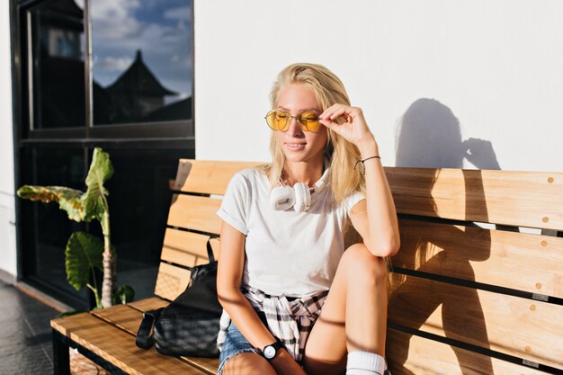 Prachtige dame in casual wit t-shirt zittend op een houten bankje. Openluchtportret van dromerige blonde vrouw die in gele zonnebril van zonnige ochtend genieten.