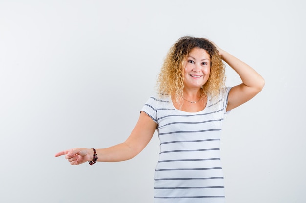 Prachtige dame in gestreept t-shirt die naar de linkerkant wijst, de andere hand op het hoofd houdt en er vrolijk uitziet, vooraanzicht.