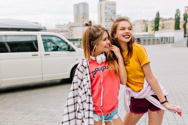 Prachtige extatische meisjes in trendy zomerkleding die tijd samen doorbrengen en genieten van uitzicht op de stad in de buurt van een witte auto