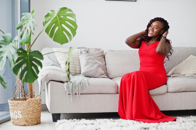 Prachtige jonge Afrikaanse vrouw in luxe rode jurk in een luxe appartement zittend op de bank Beauty fashion
