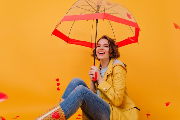 Prachtige jonge vrouw in jeans en gele jas poseren onder parasol. Indoor foto van modieus wit meisje genieten van fotoshoot in herfstdag.