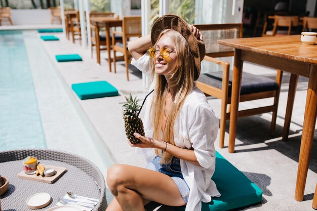 Prachtige jonge vrouw in zonnebril poseren bij zwembad met ananas. Verbazende langharige vrouwelijke toerist koelen in het resort met fruitcocktail.